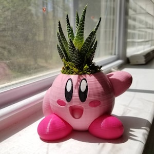May include: A pink 3D printed planter shaped like the character Kirby from the video game series Kirby. The planter has a green succulent plant growing out of the top.