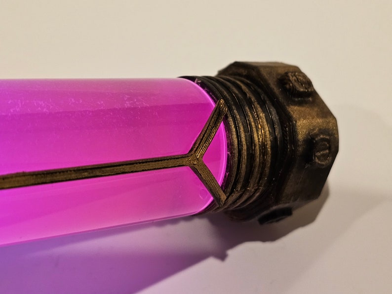 May include: A close-up of a small, cylindrical glass vial with a pink liquid inside. The vial is encased in a gold-colored metal frame with a decorative design. The vial has a metal cap with a hexagonal shape.
