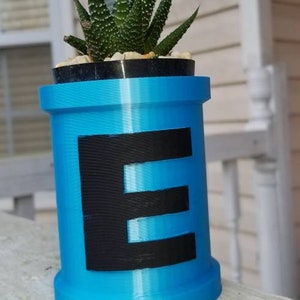 May include: A blue 3D printed planter with a black letter "E" on the front. The planter is filled with a small succulent plant.