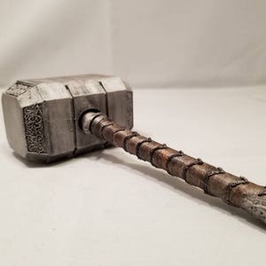 Op de afbeelding: Een zilveren en bruine hamer met een gedetailleerd ontwerp. De hamer heeft een lange steel met een gevlochten touwontwerp.
