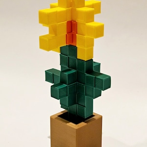 May include: A 3D printed Minecraft-style flower in a pot. The flower is yellow with an orange centre and the stem is green. The pot is brown.