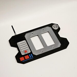 May include: A black and silver 3D printed light switch cover with two toggle switches, a red button, and a small antenna. The cover has a textured design and a blue and red stripe.