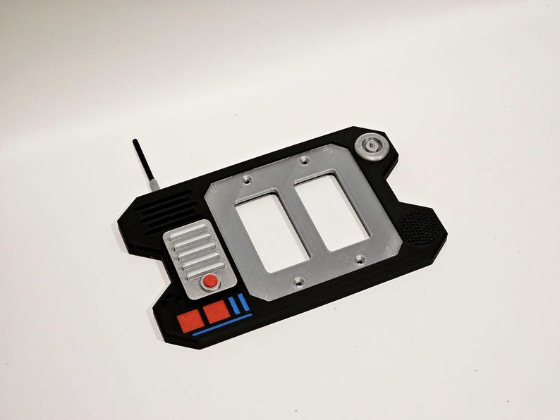 May include: A black and silver 3D printed light switch cover with two toggle switches, a red button, and a small antenna. The cover has a textured design and a blue and red stripe.
