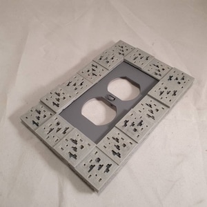 Peut inclure: Une plaque de prise de courant en plastique gris avec un motif de brique d&eacute;coratif. La plaque comporte deux prises &eacute;lectriques standard.