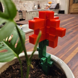May include: A red and green plastic flower with a blocky design. The flower is sitting in a white pot with soil.