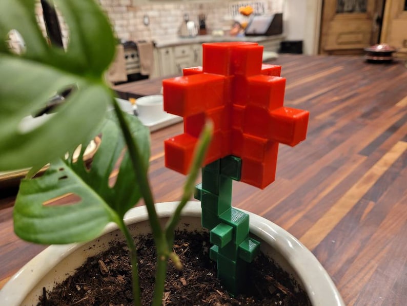 May include: A red and green plastic flower with a blocky design. The flower is sitting in a white pot with soil.