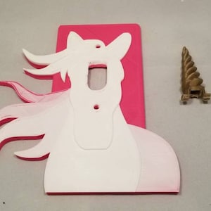 May include: A pink light switch cover with a white unicorn silhouette. The unicorn has a flowing mane and tail. A separate gold unicorn horn is included.