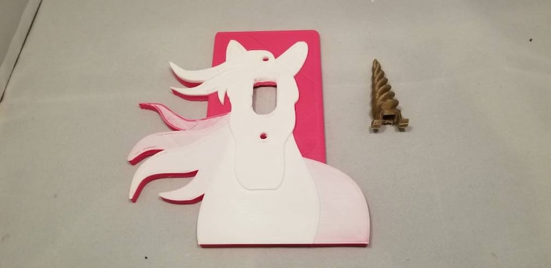 May include: A pink light switch cover with a white unicorn silhouette. The unicorn has a flowing mane and tail. A separate gold unicorn horn is included.
