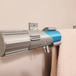 May include: A silver and blue lightsaber-shaped towel rack mounted on a white wall. The rack has a silver handle and a blue metal tube. A white towel is hanging on the rack.