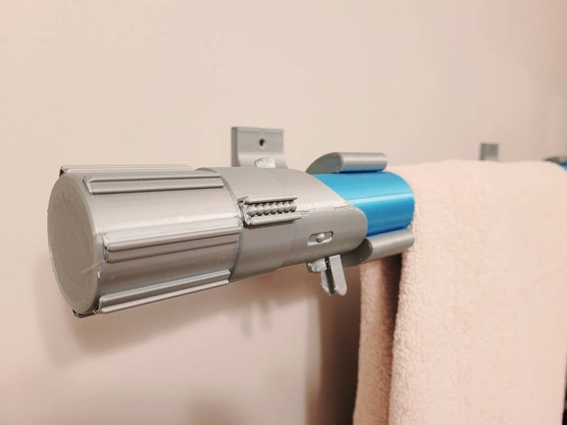May include: A silver and blue lightsaber-shaped towel rack mounted on a white wall. The rack has a silver handle and a blue metal tube. A white towel is hanging on the rack.