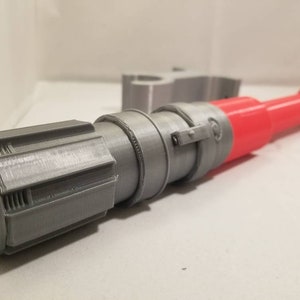 May include: A 3D-printed toy lightsaber with a red blade and gray hilt. The hilt features detailed cylindrical and rectangular components. The toy is designed to resemble a lightsaber from a science fiction franchise.