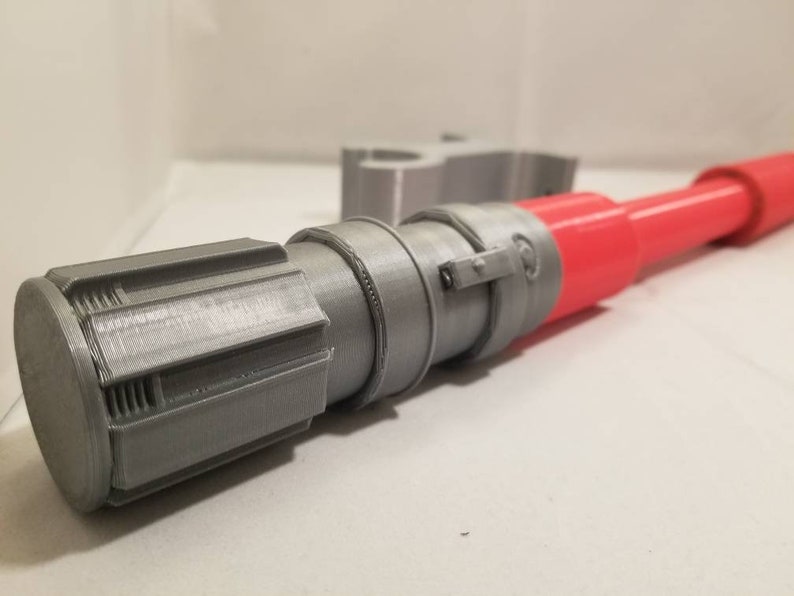 May include: A 3D-printed toy lightsaber with a red blade and gray hilt. The hilt features detailed cylindrical and rectangular components. The toy is designed to resemble a lightsaber from a science fiction franchise.