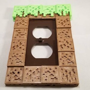 May include: A brown and green 3D printed wall plate cover with a Minecraft theme. The plate cover has a green grass block on top and brown dirt blocks on the sides. The plate cover has a standard double outlet.