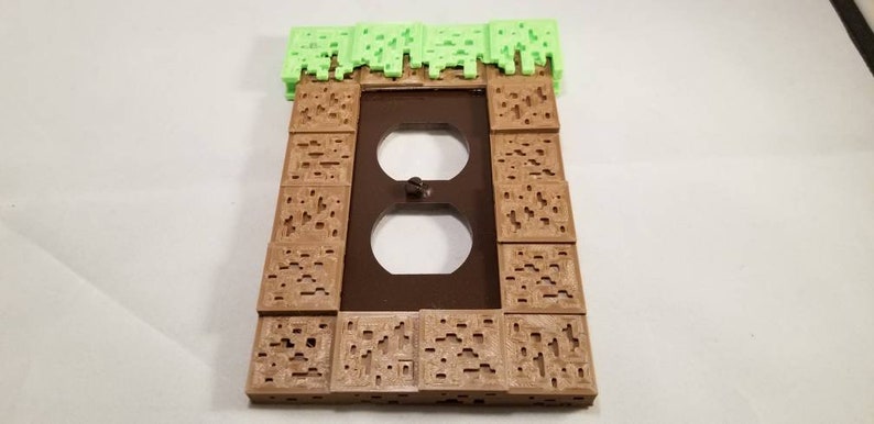 May include: A brown and green 3D printed wall plate cover with a Minecraft theme. The plate cover has a green grass block on top and brown dirt blocks on the sides. The plate cover has a standard double outlet.