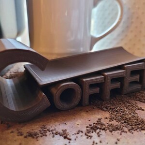May include: A brown 3D printed coffee holder with the word "COFFEE" printed on it. The holder is sitting on a surface covered in coffee grounds.