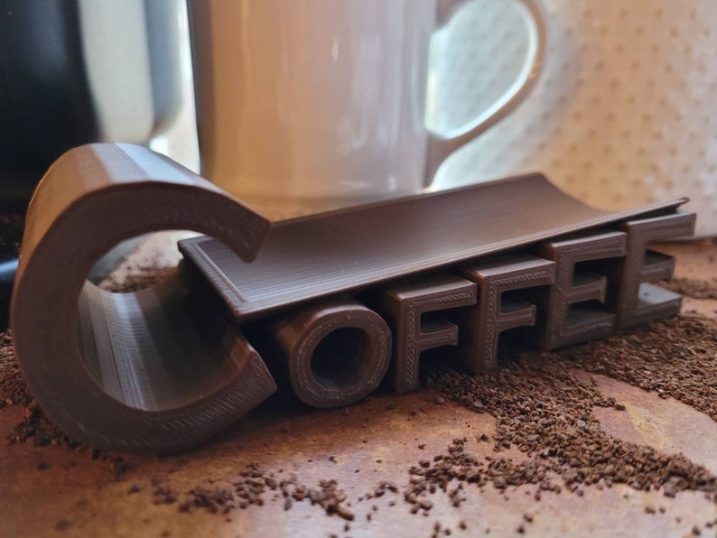 May include: A brown 3D printed coffee holder with the word "COFFEE" printed on it. The holder is sitting on a surface covered in coffee grounds.