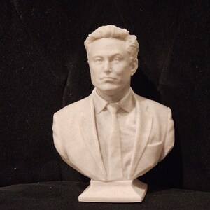 May include: A white bust sculpture of a man with short, dark hair wearing a suit jacket and tie. The bust is on a white base.