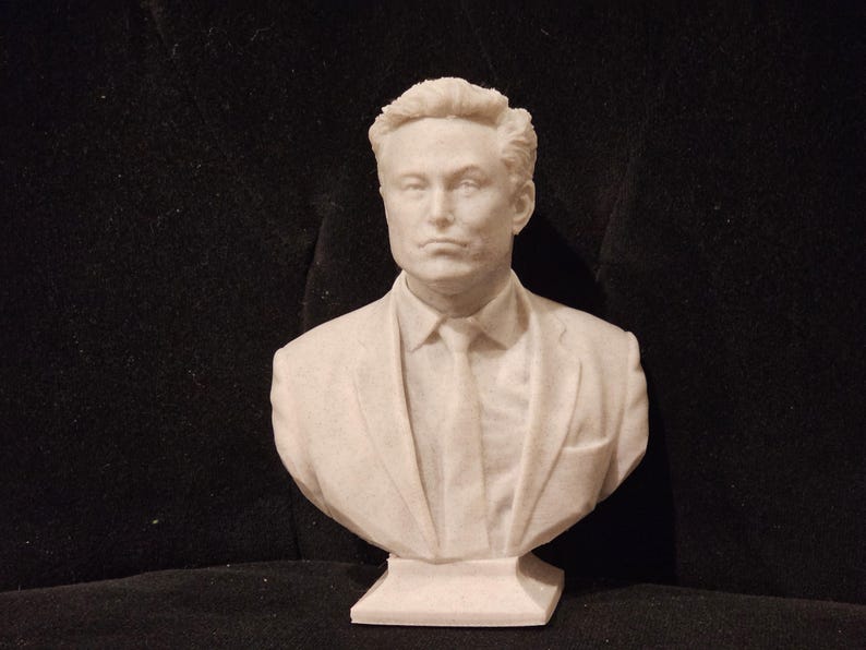 May include: A white bust sculpture of a man with short, dark hair wearing a suit jacket and tie. The bust is on a white base.