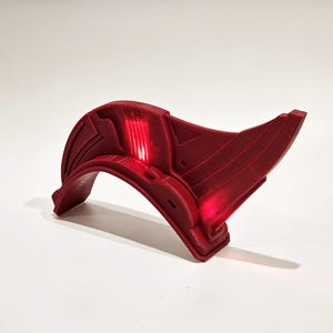 Puede incluir: Un accesorio de plástico rojo con forma curva y un extremo puntiagudo. El accesorio tiene una superficie texturizada y una luz que brilla a través de él.