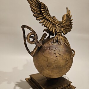 May include: A bronze-colored sculpture featuring an eagle with outstretched wings perched atop a globe. The globe rests on a stack of books. An anchor and rope detail the design. The sculpture is a decorative item.