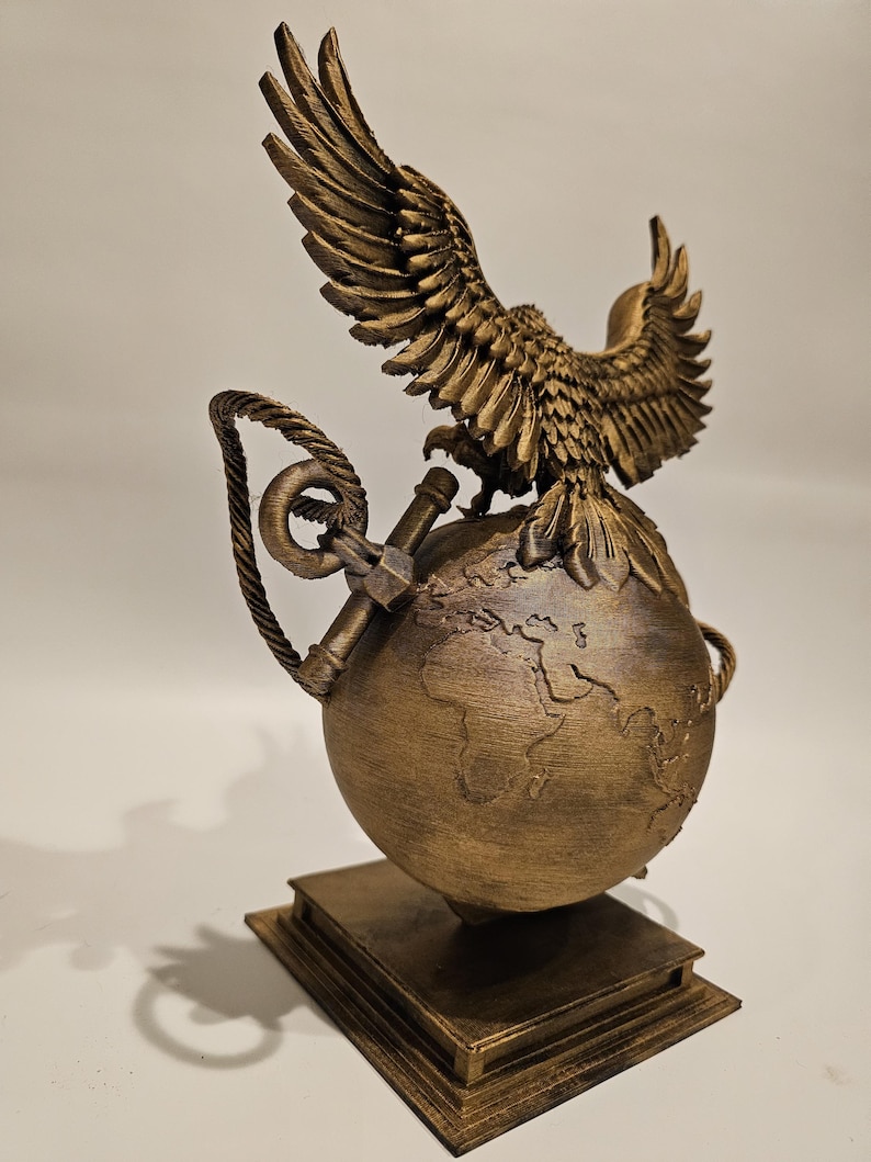 May include: A bronze-colored sculpture featuring an eagle with outstretched wings perched atop a globe. The globe rests on a stack of books. An anchor and rope detail the design. The sculpture is a decorative item.