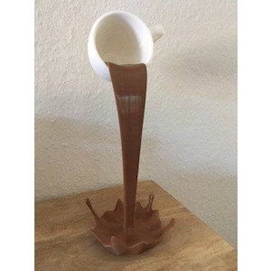 May include: A white ceramic cup pouring brown liquid into a brown, splatter-shaped base.