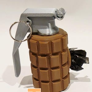 May include: A tan-colored grenade with a silver-colored top and pull ring. The body of the grenade has a textured, block-like design. The word "Tan" is written in orange at the bottom of the image.