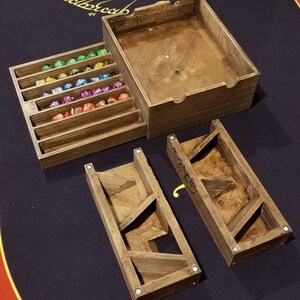 May include: A wooden dice tower with a separate compartment for storing dice. The tower has a sloped design with multiple levels for the dice to roll down. The dice tower is made of natural wood with a light brown finish. The dice tower is sitting on a red and black felt gaming table.