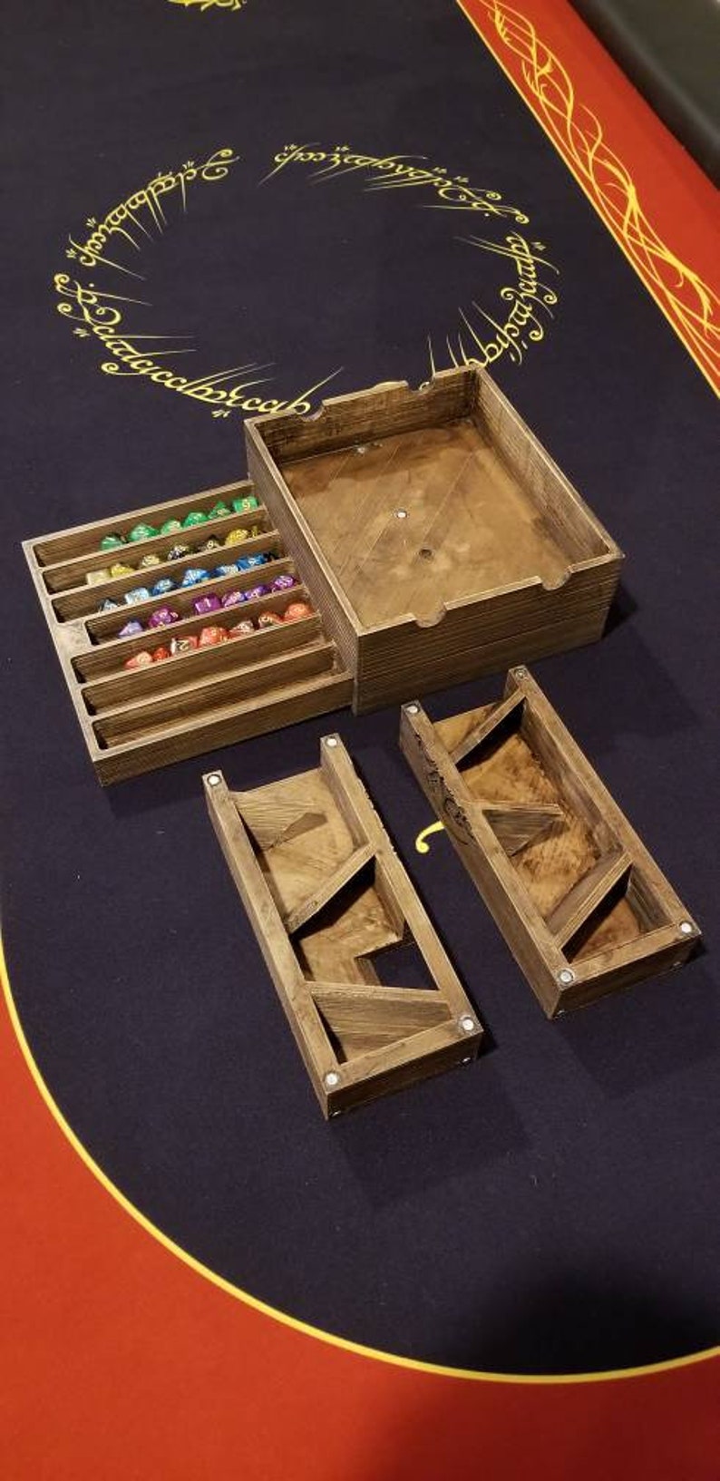 May include: A wooden dice tower with a separate compartment for storing dice. The tower has a sloped design with multiple levels for the dice to roll down. The dice tower is made of natural wood with a light brown finish. The dice tower is sitting on a red and black felt gaming table.