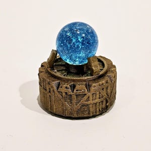 May include: A bright blue crackle glass sphere sits atop a circular, bronze-coloured base with a textured, architectural design. The base features vertical ridges and a central platform held by small supports, creating a detailed, layered appearance.