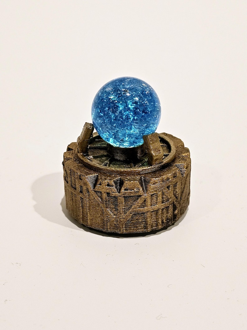 May include: A bright blue crackle glass sphere sits atop a circular, bronze-coloured base with a textured, architectural design. The base features vertical ridges and a central platform held by small supports, creating a detailed, layered appearance.