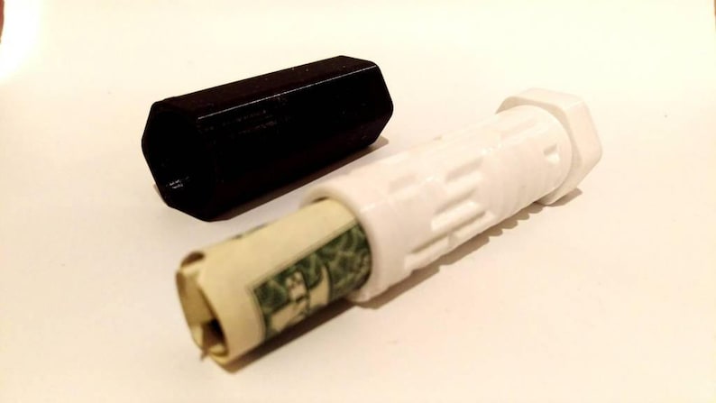 May include: A white plastic tube with a black cap. The tube is designed to hold a rolled up dollar bill. The tube has a textured surface and a hexagonal shape.