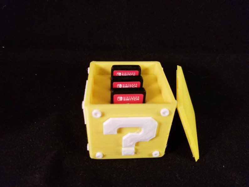 K&ouml;nnte beinhalten: Eine gelbe Plastikbox in Form eines Fragezeichenblocks aus dem Videospiel Super Mario Bros. Die Box hat einen Klappdeckel und fasst drei Nintendo Switch-Spielkassetten. Die Kassetten sind mit "Nintendo Switch" beschriftet.