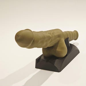 May include: A tan-colored sculpture of a cannon with a phallic design. The cannon is mounted on a black base. The sculpture has detailed textures and is set against a plain white background.