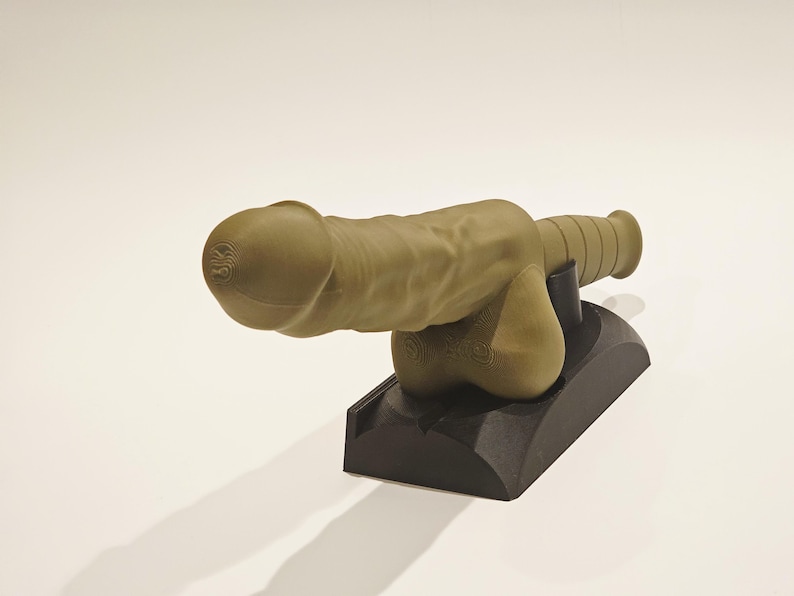 May include: A tan-colored sculpture of a cannon with a phallic design. The cannon is mounted on a black base. The sculpture has detailed textures and is set against a plain white background.