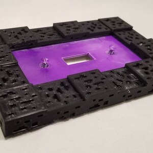May include: A rectangular black frame with a textured, geometric design. The frame surrounds a vibrant purple surface with a rectangular cutout and two silver screws. The overall design is modern and abstract.