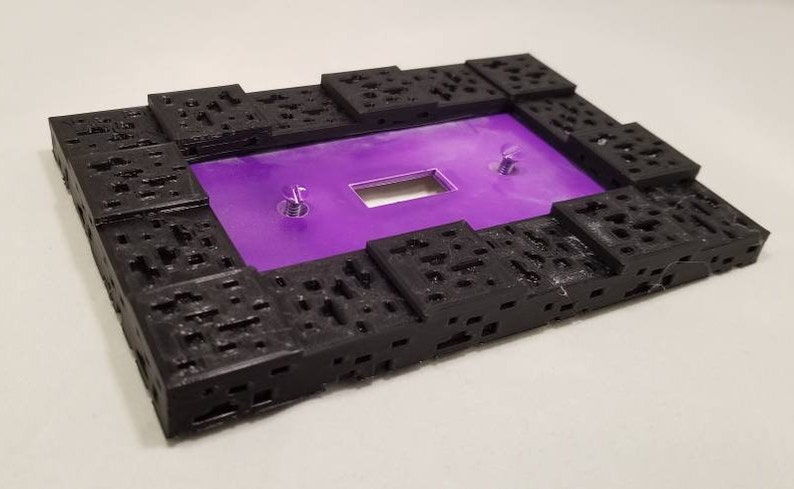 May include: A rectangular black frame with a textured, geometric design. The frame surrounds a vibrant purple surface with a rectangular cutout and two silver screws. The overall design is modern and abstract.