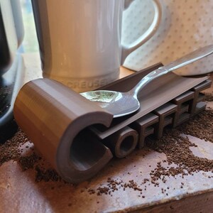 May include: A brown 3D printed coffee spoon rest with the word "COFFEE" printed on it. A silver spoon is resting on the rest.