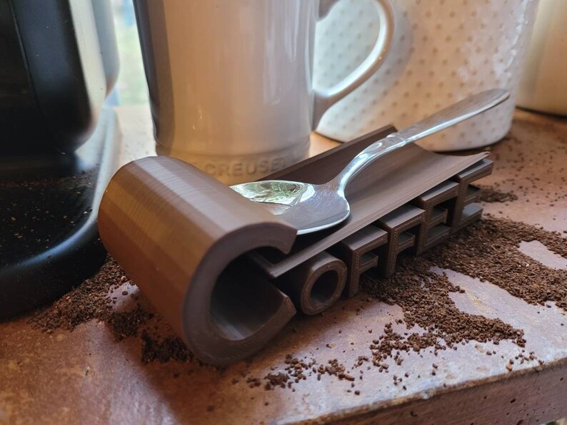 May include: A brown 3D printed coffee spoon rest with the word "COFFEE" printed on it. A silver spoon is resting on the rest.