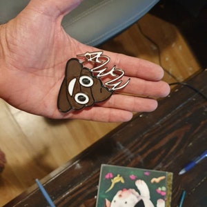 May include: A brown and white poop emoji keychain with the word "Poop" written in white on top. The keychain is being held in a person's hand.