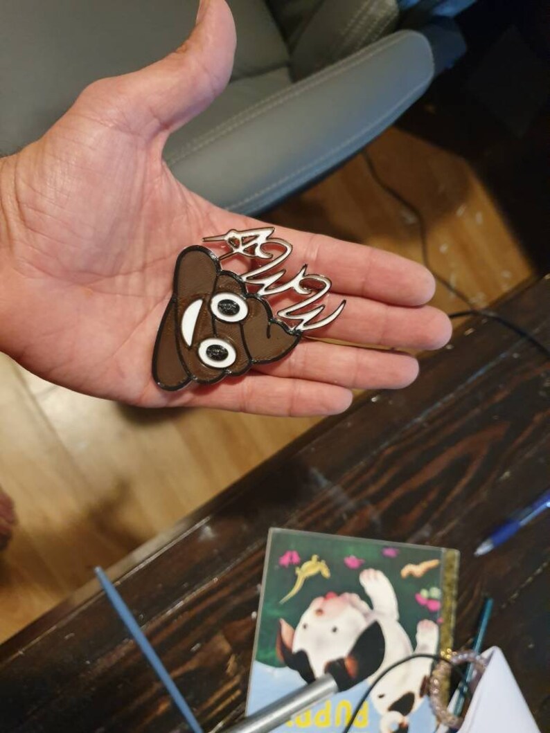 May include: A brown and white poop emoji keychain with the word "Poop" written in white on top. The keychain is being held in a person's hand.