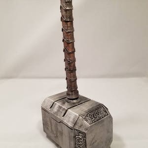 Op de afbeelding: Een zilverkleurige Mjolnir-hamervormige doos met een bruine houten handgreep. De doos heeft ingewikkelde details en een getextureerd oppervlak.