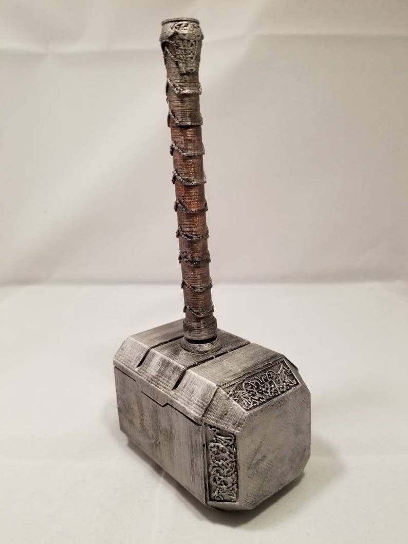 Op de afbeelding: Een zilverkleurige Mjolnir-hamervormige doos met een bruine houten handgreep. De doos heeft ingewikkelde details en een getextureerd oppervlak.