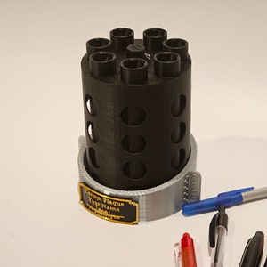May include: A black 3D printed pen holder with a gray base and a custom plaque that says "Custom Plaque Your Name". The holder has eight slots for pens and is sitting on a white surface with several pens next to it.