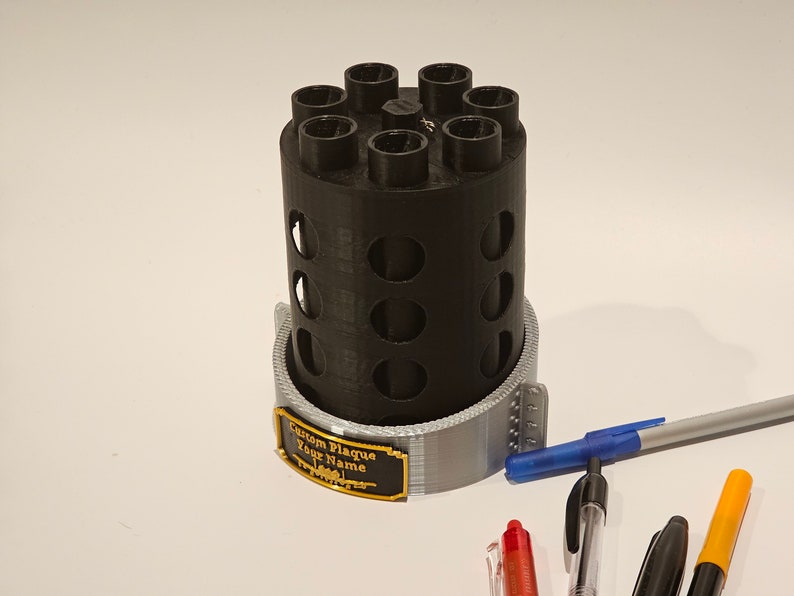 May include: A black 3D printed pen holder with a gray base and a custom plaque that says "Custom Plaque Your Name". The holder has eight slots for pens and is sitting on a white surface with several pens next to it.