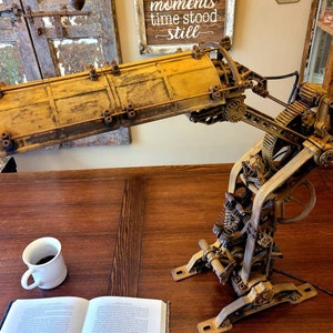 Puede incluir: Una l&aacute;mpara de estilo steampunk con una pantalla de metal amarilla y engranajes y mecanismos intrincados. La l&aacute;mpara est&aacute; sobre una mesa de madera con un libro y una taza de caf&eacute;.