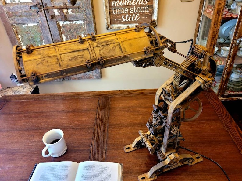 Puede incluir: Una l&aacute;mpara de estilo steampunk con una pantalla de metal amarilla y engranajes y mecanismos intrincados. La l&aacute;mpara est&aacute; sobre una mesa de madera con un libro y una taza de caf&eacute;.