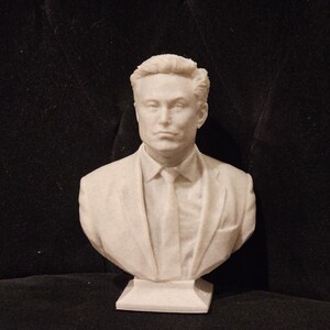 May include: A white bust sculpture of a person with short brown hair wearing a suit jacket and tie. The bust is on a white base.