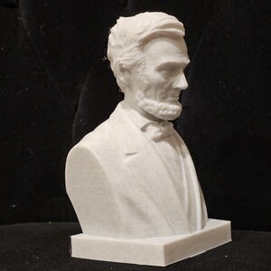 May include: White plaster bust of Abraham Lincoln, a historical figure, with a detailed face and a suit jacket. The bust is on a white base.