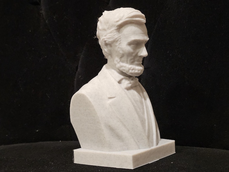 May include: White plaster bust of Abraham Lincoln, a historical figure, with a detailed face and a suit jacket. The bust is on a white base.